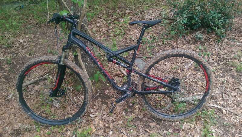 A black mountain bike with red accents resting on the ground, surrounded by foliage and fallen leaves. The bike appears to be muddy, indicating recent use on a dirt trail. Sope Creek mountain bike trail.