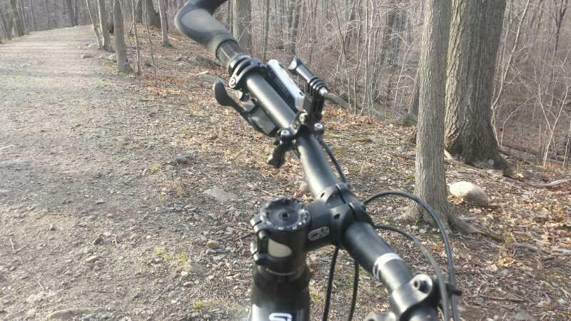 A close-up view of a mountain bike handlebar, showing the control grips and gears, with a dirt path winding through a wooded area in the background. The trees are bare, suggesting early spring or late autumn. Tourne Park mountain bike trail.