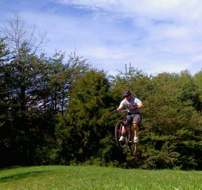 A person on a mountain bike performing a jump over a grassy area, surrounded by trees under a blue sky. Sherando Park Mountain Bike Trails mountain bike trail.