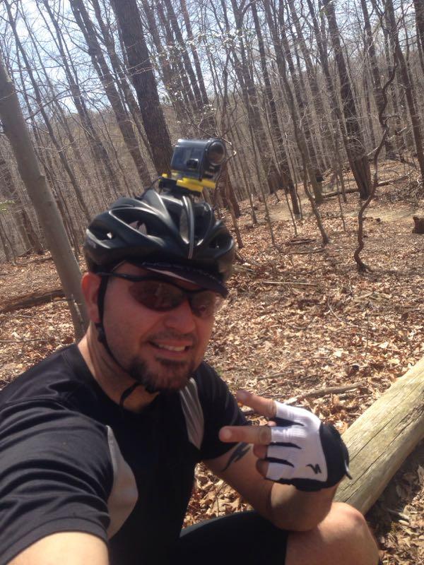 A person wearing a black cycling helmet and sunglasses smiles while taking a selfie in a wooded area. They are dressed in a short-sleeved cycling shirt and fingerless gloves, and there is a camera mounted on their helmet. The ground is covered in leaves, and trees surround the scene, suggesting an outdoor biking adventure. Fountainhead Regional Park mountain bike trail.