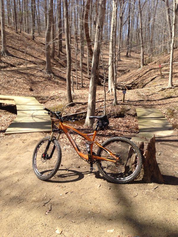 An orange mountain bike parked on a dirt trail next to a wooden bridge in a forested area with bare trees and fallen leaves on the ground. Fountainhead Regional Park mountain bike trail.