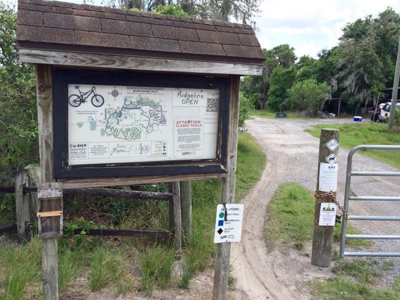 A wooden information sign displaying a map of bike trails, with the text "Ridgeline OPEN" handwritten at the top. Below the map, an announcement states "ATTENTION: CLOSED TRAILS." A gravel path leads off to the right, next to a gate marked with various trail symbols and additional signs. Surrounding vegetation includes grass and small shrubs. Balm Boyette Scrub Preserve mountain bike trail.