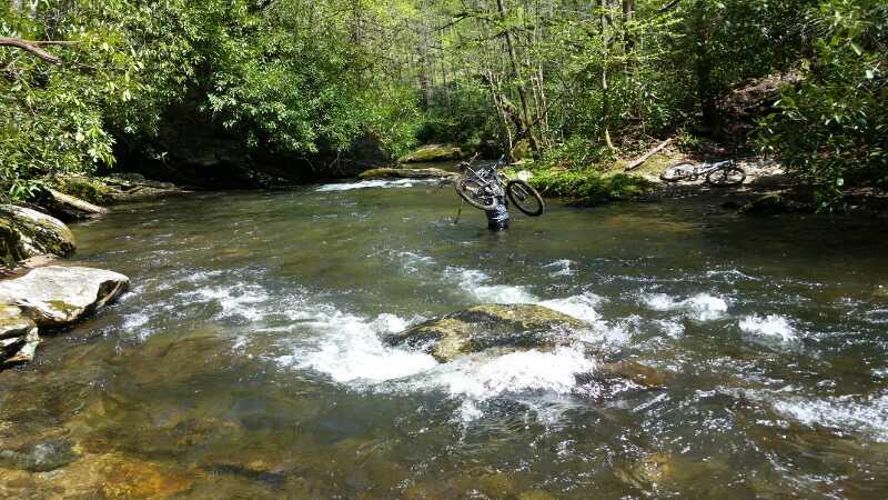 A cyclist wades through a clear, flowing river, holding their bike above the water. Lush green trees surround the riverbank, creating a serene and natural setting. Another bike is partially visible on the riverbank. Sunlight filters through the foliage, illuminating the scene. South Mills River Trail mountain bike trail.