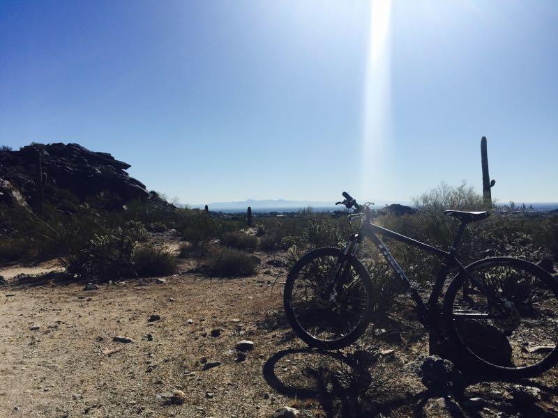 Mountain bike resting on a sandy trail with desert vegetation and rock formations in the background, under a clear blue sky. Sunlight casts a bright beam over the scene, highlighting the mountain range in the distance. Desert Classic mountain bike trail.