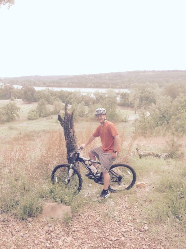 A person wearing a helmet and an orange shirt stands next to a mountain bike on a grassy hillside. In the background, there is a view of a body of water and trees, indicating a natural outdoor setting. Burns Park mountain bike trail.