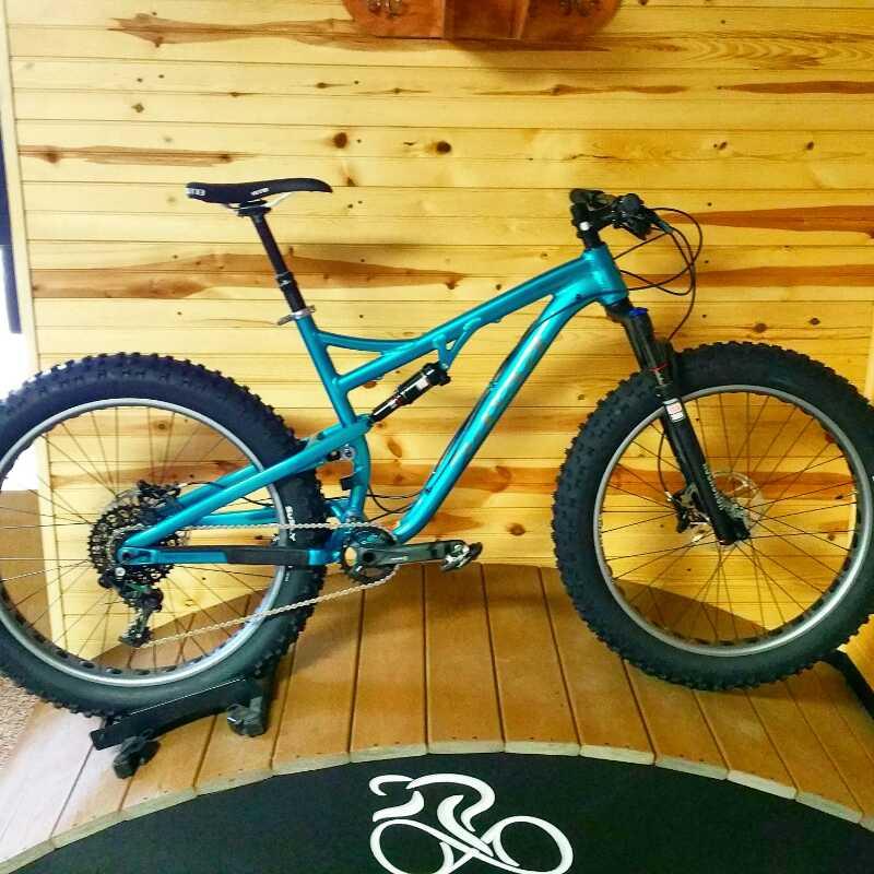 A bright blue mountain bike with thick tires is displayed on a wooden floor, set against a background of wooden wall panels. The bike features a suspension system and a sleek frame design, resting on a bike stand in front of a circular mat with a bicycle graphic.