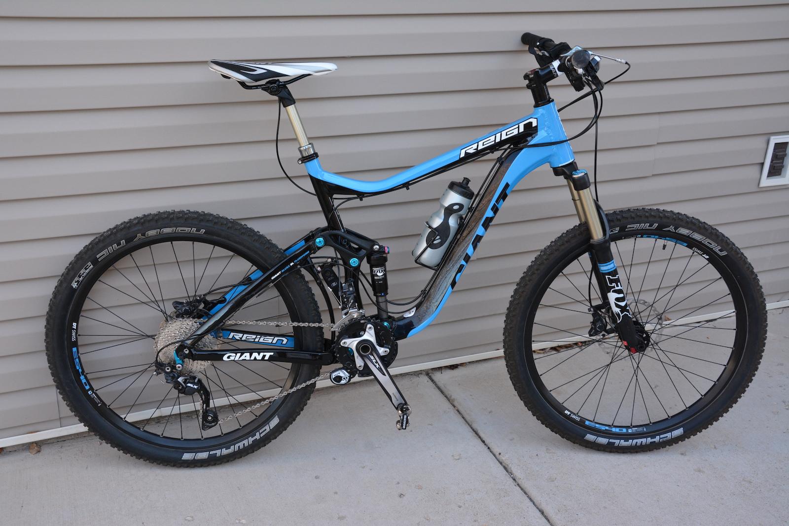 Giant Reign: A blue and black mountain bike leaning against a beige wall. The bike features a full suspension frame, disc brakes, and a water bottle holder. The tires are designed for off-road terrain, displaying a rugged tread pattern. The bike is positioned on a concrete surface, highlighting its sleek design.