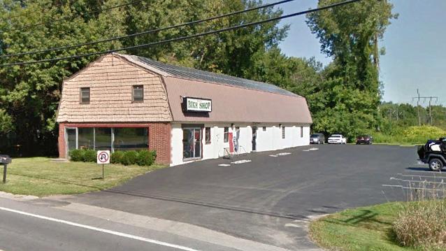 A brick shop building with a sloped roof, featuring large windows and a sign that reads "Bike Shop." The parking lot in front is paved, and there are a few parked cars. Surrounding the building are trees and green space, indicating a rural or suburban setting.