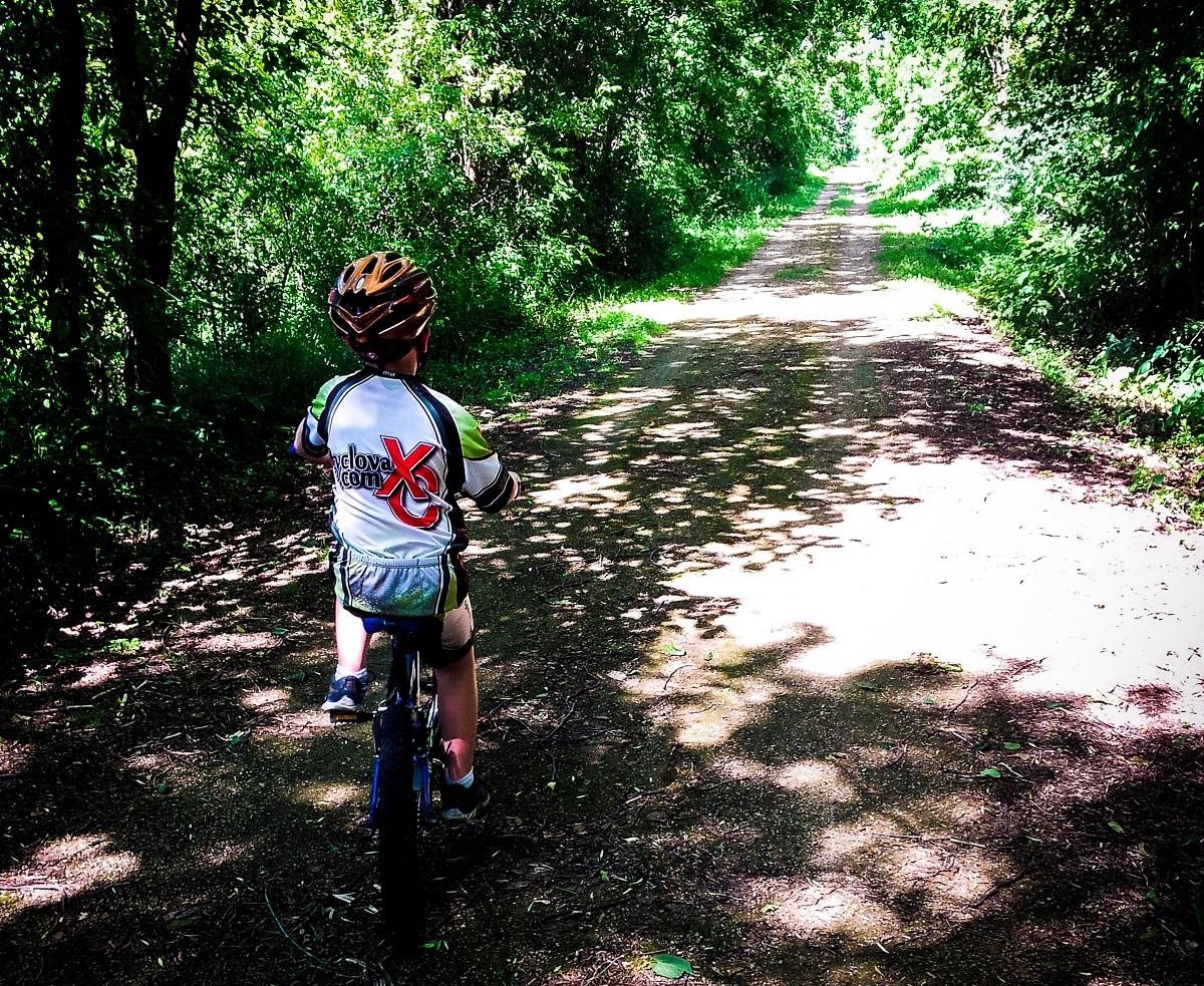 A child in a cycling helmet rides a small bike along a dirt path surrounded by lush greenery and dappled sunlight. The path stretches ahead through a serene wooded area. Gandy Dancer mountain bike trail.