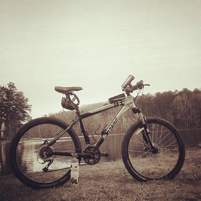 Scott Reflex 20: A mountain bike parked on grassy terrain near a calm lake, surrounded by trees and hills, with a sepia tone effect enhancing the serene atmosphere.