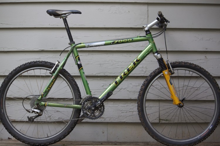 Alt text: A green Trek mountain bike stands against a textured gray wall. The bike features a front suspension fork with an orange accent, a black saddle, and knobby tires, indicating it is designed for off-road riding.