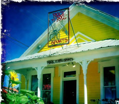 A bright yellow building with a decorative sign reading "Rock and Roll Sports," featuring a bicycle graphic. The storefront has white trim, and there is a sunflower in the foreground. The entrance is open, indicating business hours.