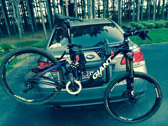 Giant Trance X2: A blue mountain bike mounted on the back of a silver car in a wooded area. The bike features the brand name "Giant" prominently displayed on the frame and wheels. The background shows tall trees, suggesting a natural setting.
