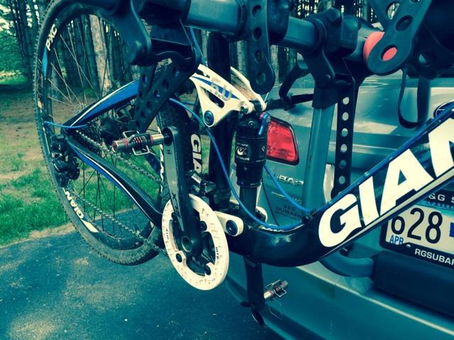 Motobecane NightTrain Bullet: A close-up view of a blue Giant mountain bike mounted on a bike rack, showcasing the rear suspension and drivetrain components. The background features a silver vehicle and a wooded area. The bike has visible dirt, indicating recent use.