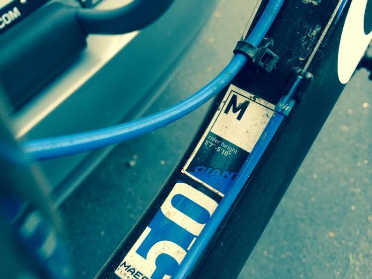 Motobecane NightTrain Bullet: Image of a bicycle frame displaying a size label indicating "M" for medium, along with a rider height range of 5'7" to 5'10". Blue cables are visible alongside the label, suggesting it is part of a bike's gear system. The background is slightly blurred, revealing a glimpse of a vehicle.