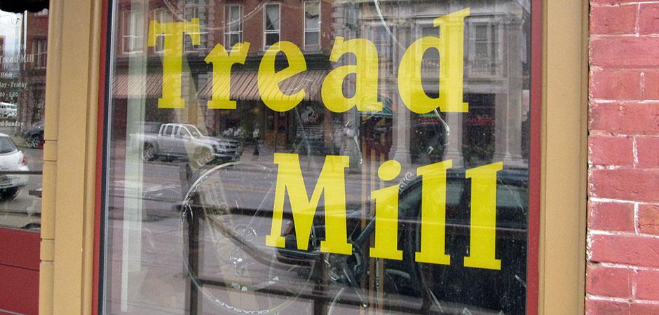Sign on a storefront window displaying the words "Tread Mill" in large yellow letters. The background shows a reflection of buildings and vehicles on a street.