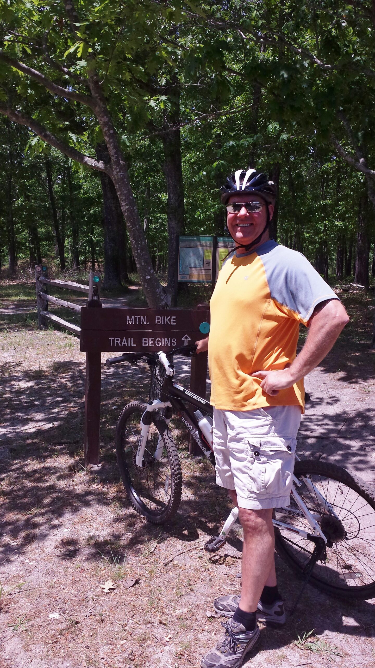 A man wearing a helmet and sunglasses stands next to a mountain bike at a trailhead marked "MTN. BIKE TRAIL BEGINS." The scene is surrounded by lush greenery and a mix of trees in a sunny outdoor setting. Wharton State Forest mountain bike trail.