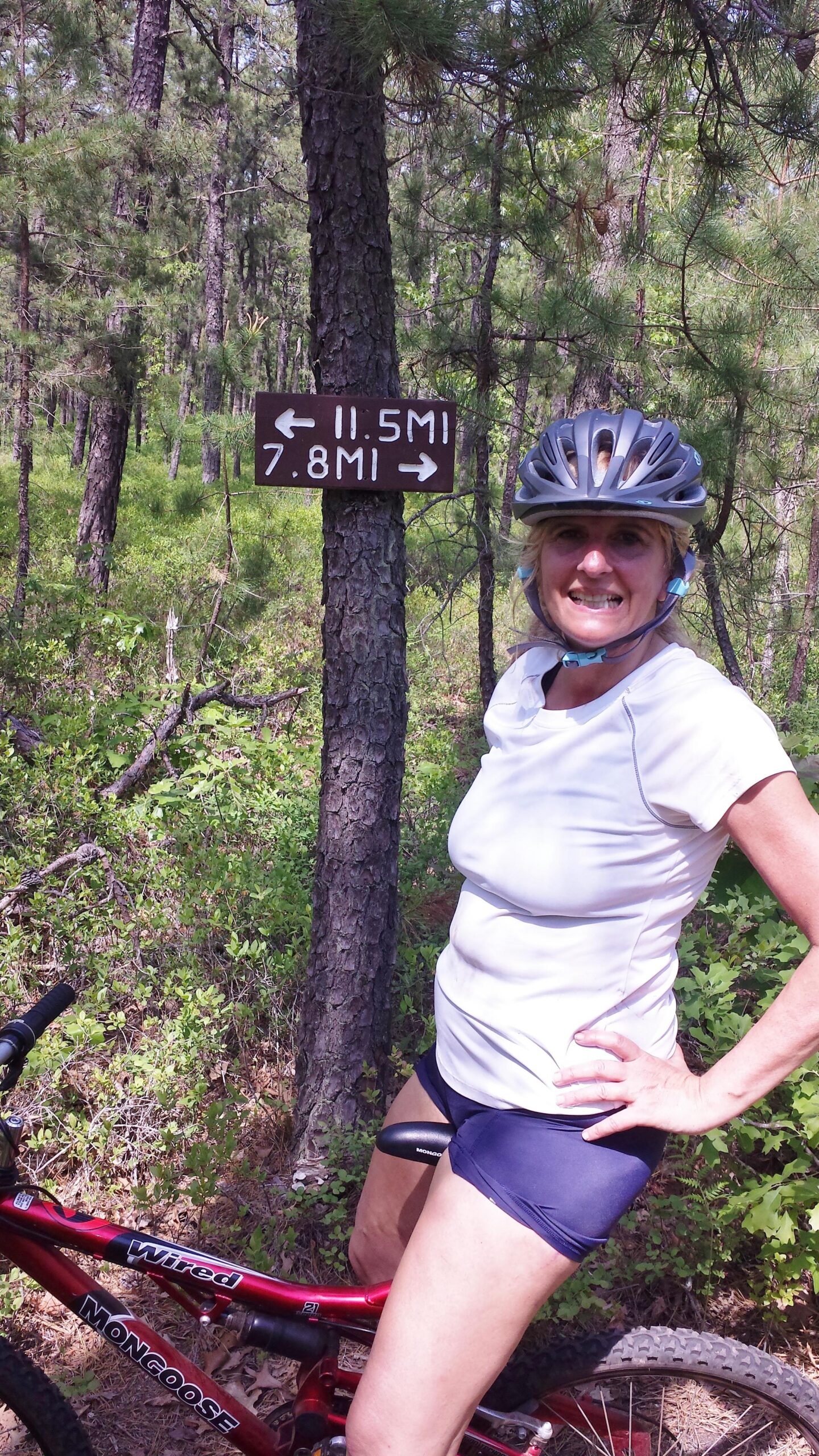 A smiling woman in a white athletic shirt and bike shorts stands next to a trail sign in a forested area. The sign indicates distances of 11.5 miles and 7.8 miles in both directions. She is wearing a bicycle helmet and has her hands on her hips, with a red mountain bike beside her. Wharton State Forest mountain bike trail.