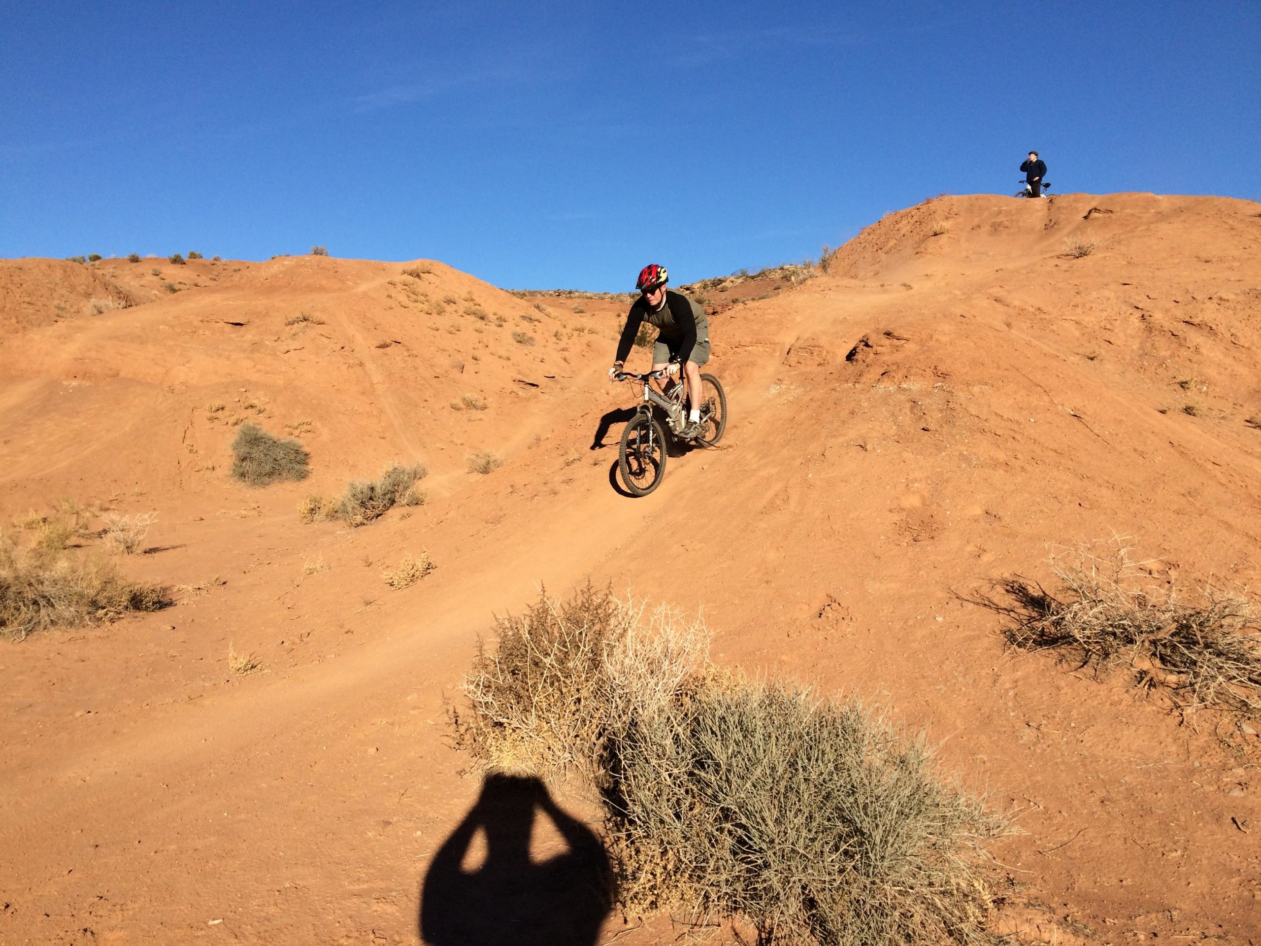 A mountain biker riding down a dirt hill in a desert landscape, with another biker visible on a higher vantage point in the background. The scene features sandy terrain, sparse vegetation, and a clear blue sky. Bearclaw Poppy mountain bike trail.