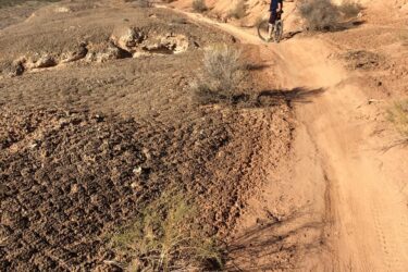 A cyclist riding on a dirt trail surrounded by rocky terrain, with a mountainous backdrop under a clear blue sky. The path winds along the base of a cliff, showcasing the natural landscape and dry vegetation typical of a desert environment. Bearclaw Poppy mountain bike trail.