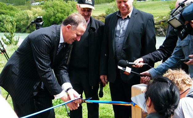 A man in a suit is cutting a ribbon with scissors as part of a ceremonial event, surrounded by a small group of onlookers and media. The background features a river and green hills. The scene appears to be an official inauguration or opening celebration. The New Zealand Cycle Trail mountain bike trail.