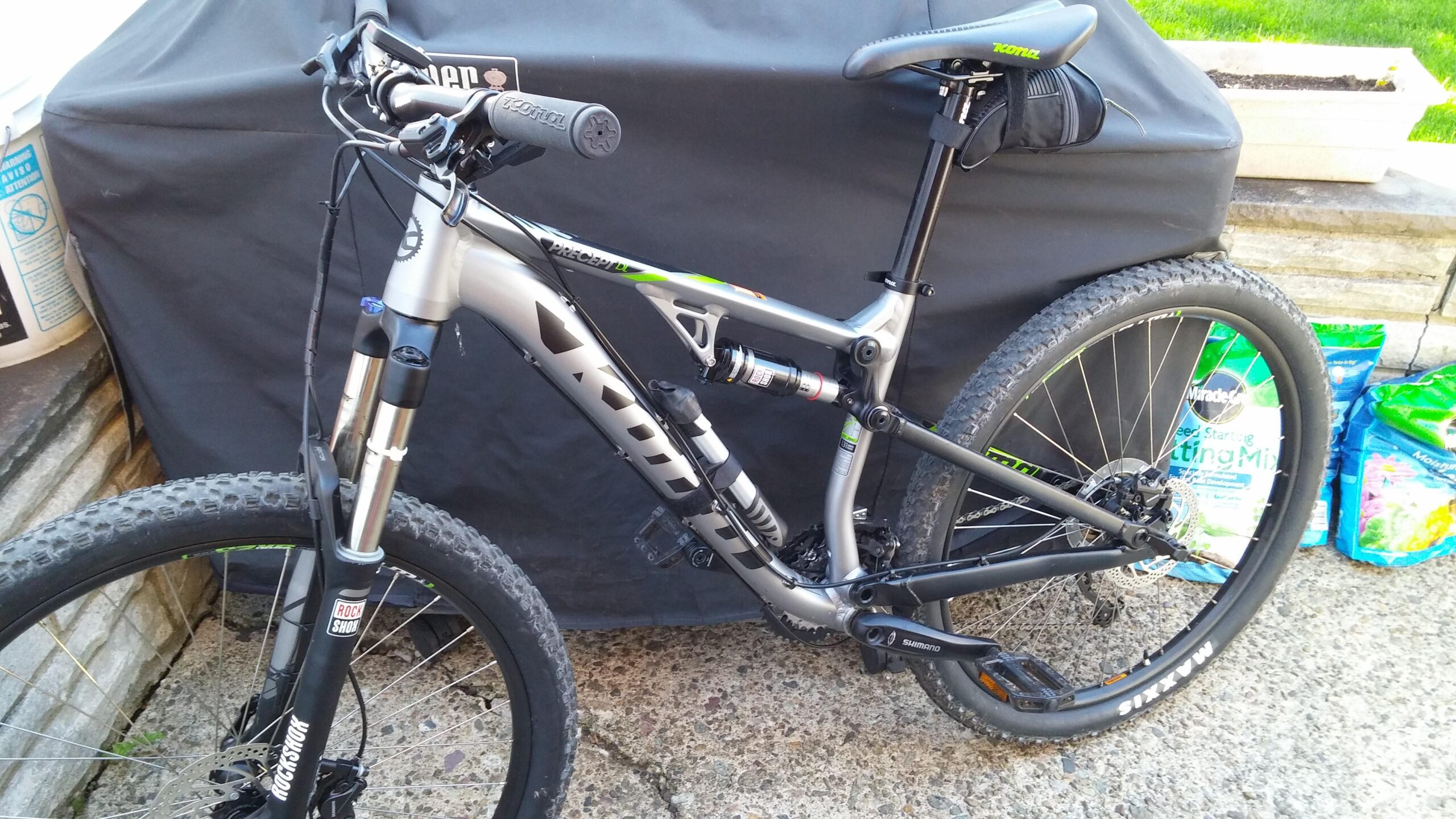 Kona Precept DL: A mountain bike with a sleek silver and black frame, featuring front suspension with RockShox components and disc brakes. The bike is positioned against a dark cover and surrounded by bags of planting mix. The scene includes hints of a garden or outdoor area.