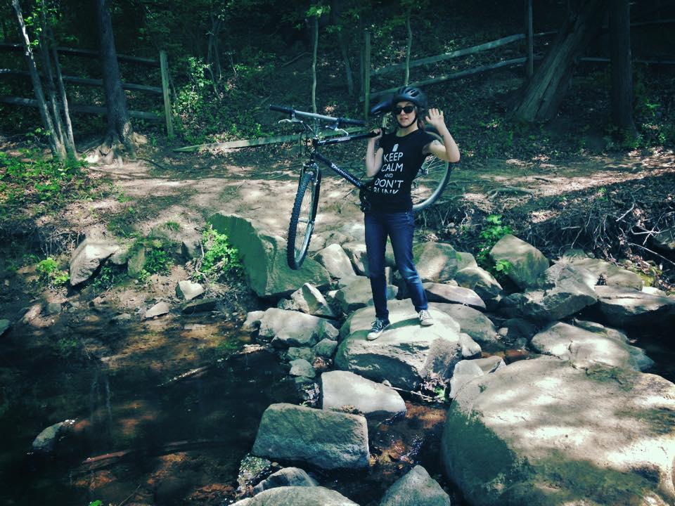 A person standing on large rocks near a stream, holding a bicycle over their shoulder. They are wearing a black tank top with a slogan, sunglasses, and jeans. The surrounding area is lush with greenery and trees, indicating a natural outdoor setting. Laurel Hill Park mountain bike trail.