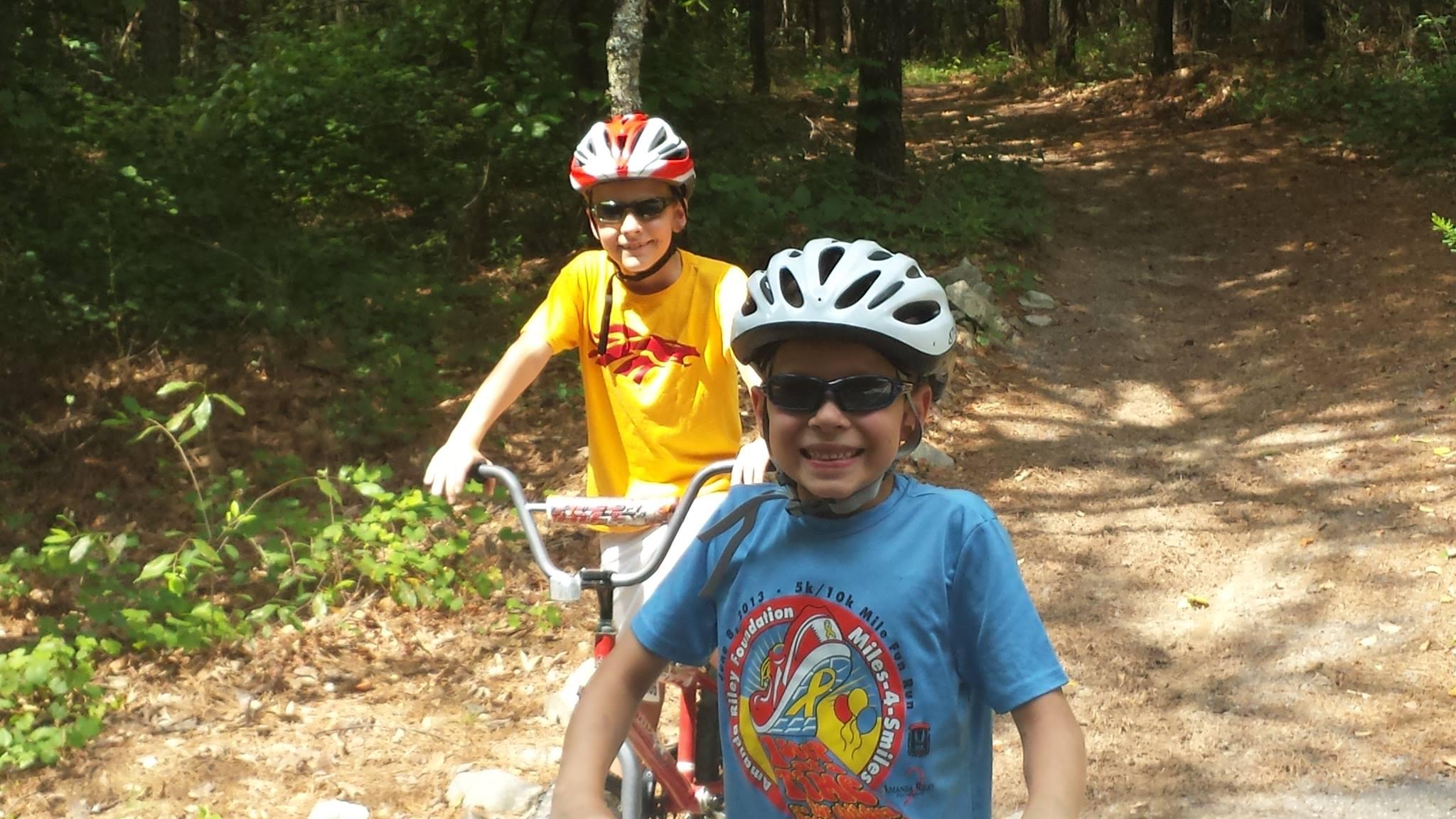 Two children wearing helmets and sunglasses are smiling on a dirt biking trail surrounded by greenery. One child is riding a bicycle while the other is standing beside it, both looking cheerful and ready for an adventure. The scene captures a sunny day in a forested area. Harbins Park mountain bike trail.