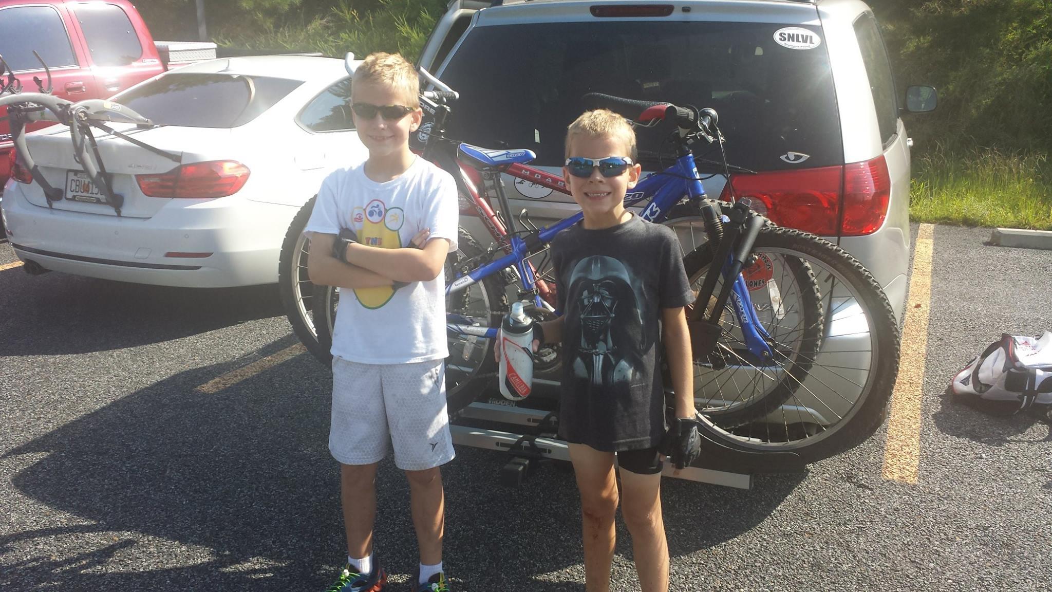 Two young boys stand outdoors beside a vehicle in a parking area. One boy is wearing a white t-shirt with a colorful design and shorts, while the other is wearing a black t-shirt featuring a character design and shorts. Both are wearing sunglasses and smiling. Behind them, a blue mountain bike is mounted on the back of the vehicle. The scene appears sunny and casual, suggesting a day of outdoor activities. Harbins Park mountain bike trail.