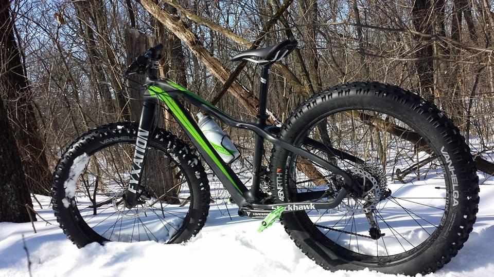 Fyxation Fyxation Blackhawk: A black and green fat bike parked in a snowy forest, with large tires designed for winter conditions. The bike is positioned against a backdrop of bare trees and scattered snow, showcasing its rugged design and sturdy frame.