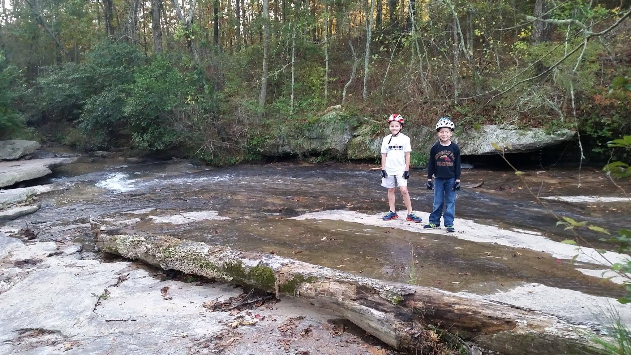 Two children stand on a rocky riverbank, surrounded by trees and greenery. They are wearing helmets and casual clothing, with a small stream flowing past them. A fallen log is visible on the ground, and autumn leaves are scattered around. The scene captures a moment of outdoor adventure in a natural setting. Harbins Park mountain bike trail.