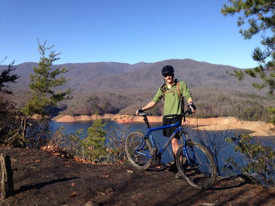 A cyclist stands next to a blue mountain bike on a rocky overlook with a scenic view of a lake and mountains in the background. The sky is clear and blue, and trees are visible in the foreground, hinting at a natural, outdoor setting. The cyclist is wearing a helmet and a green long-sleeve shirt, smiling as they pose for the photo. Tsali Recreation Area mountain bike trail.