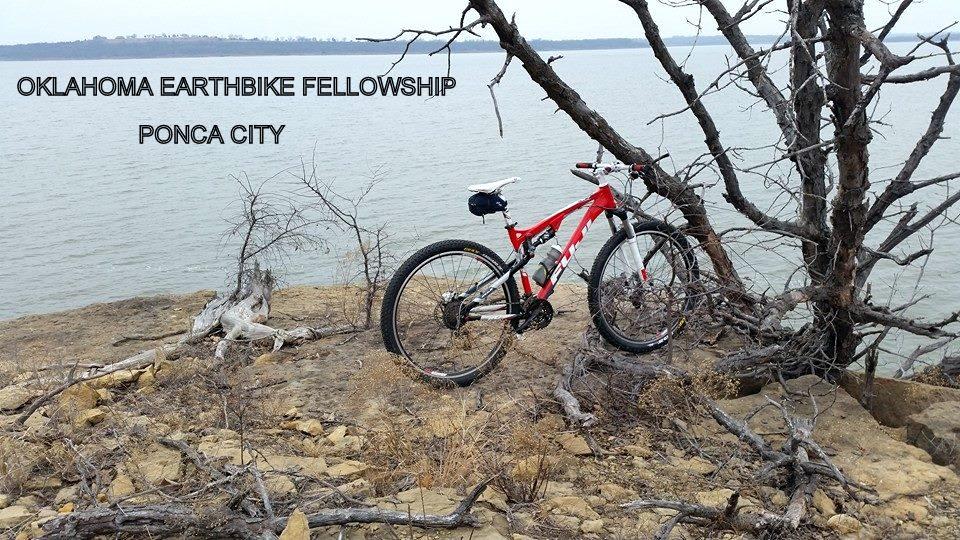 A red mountain bike parked on a rocky shoreline with a calm body of water in the background, surrounded by bare trees and brush. The text overlay reads "OKLAHOMA EARTHBIKE FELLOWSHIP PONCA CITY."