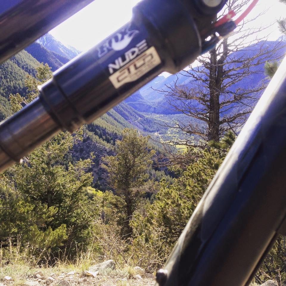 A close-up view of a bicycle shock absorber with a mountainous landscape in the background, showcasing pine trees and rolling hills under a clear sky. Colorado Trail: Mt. Shavano thd to Chalk Creek thd mountain bike trail.