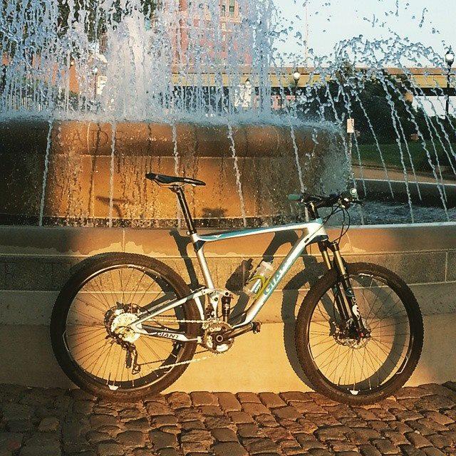 Giant Anthem: A mountain bike leaning against a stone fountain, with water cascading down in the background. The bike has a silver and blue frame, and the sun is casting a warm glow on the scene, highlighting the texture of the cobblestone ground.