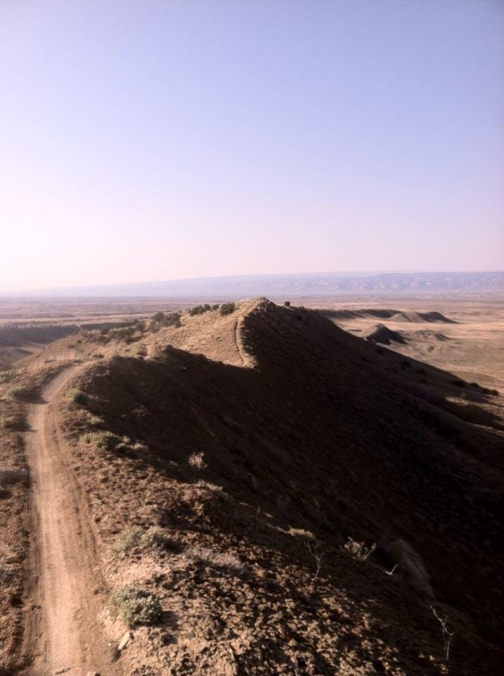 A panoramic view of a rugged landscape featuring rolling hills under a clear blue sky. A dirt trail winds through the foreground, leading up a prominent hilltop. The terrain is dry with sparse vegetation, and the horizon stretches out to reveal distant hills and valleys. The scene conveys a sense of tranquility and natural beauty. Zippety Do Dah mountain bike trail.