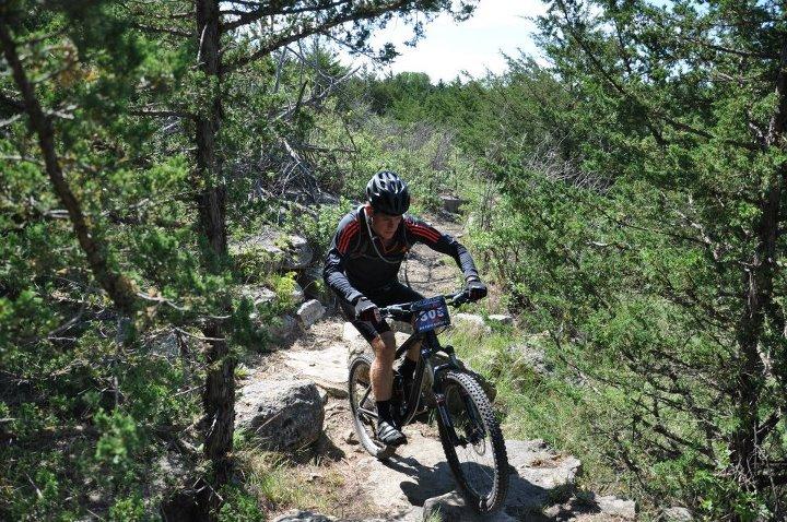 Trek Fuel EX 8: A mountain biker navigating a rocky trail surrounded by greenery on a sunny day. The rider is wearing a helmet and athletic gear, focused on the path ahead.
