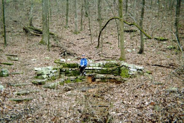 A person wearing a blue jacket sits on a stone foundation in a wooded area, surrounded by trees and fallen leaves. The remnants of the structure are partially covered in moss, and the forest floor is covered with dried leaves and scattered stones. Bench Trail mountain bike trail.
