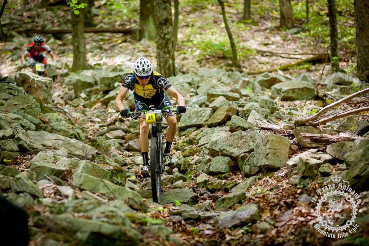 A mountain biker navigating a rocky trail in a forested area, with lush green trees in the background. The cyclist, wearing a black and yellow jersey and a helmet, is focused on maneuvering through the challenging terrain. Another rider can be seen in the background, also tackling the rocky path.
