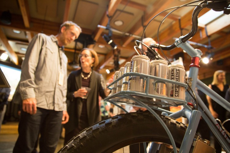 A close-up view of a bicycle with a rack holding several cans, with two people in conversation in the background. The setting appears to be a lively event indoors, featuring wooden beams and warm lighting.