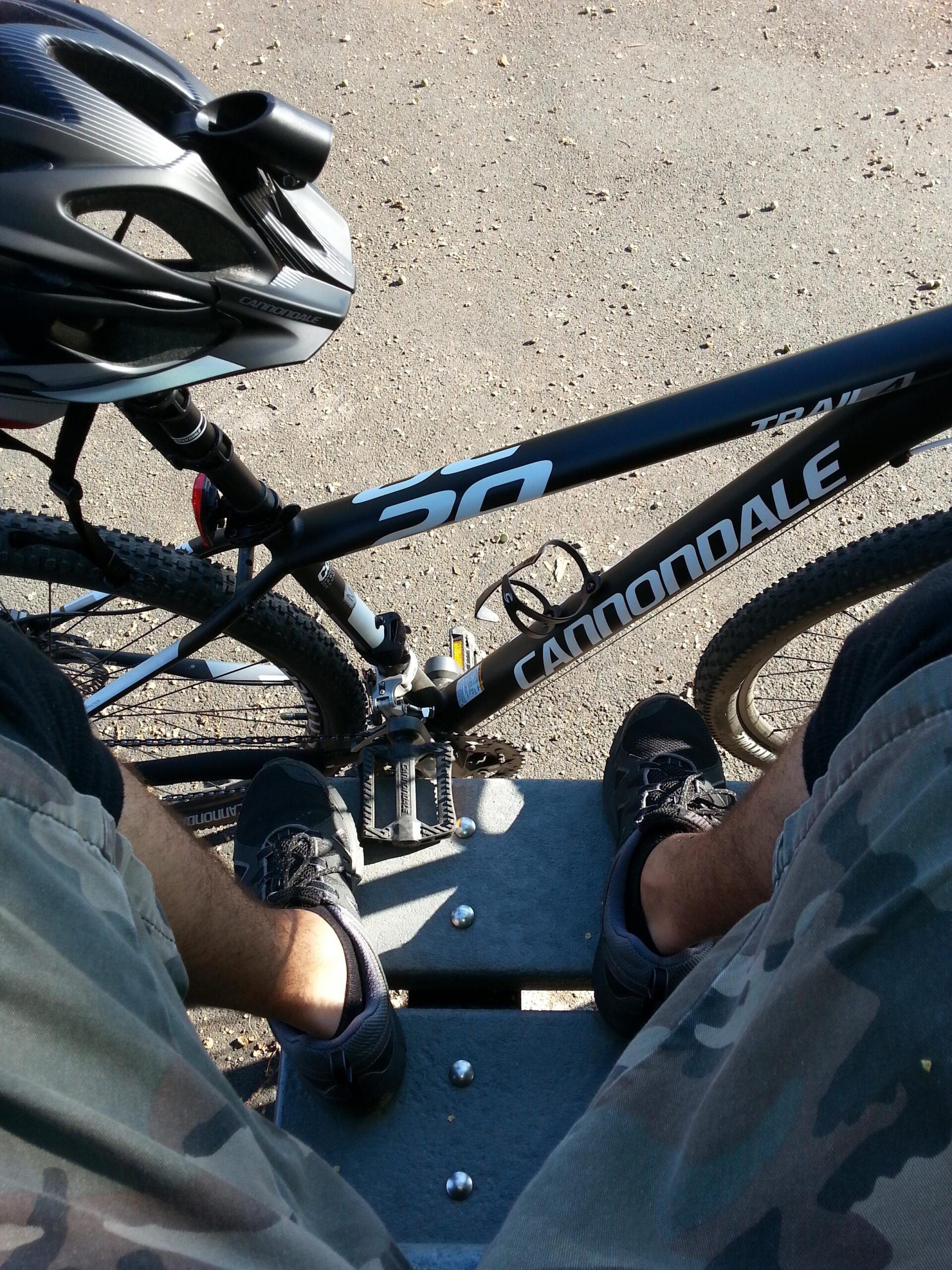 Cannondale Trail 29 4: Aerial view of a partially visible mountain bike resting on a bench, with the rider's legs in camouflage shorts and black shoes. A black bicycle helmet is positioned on the bike, surrounded by a gravel path. Sunlight highlights the scene, suggesting an outdoor setting.