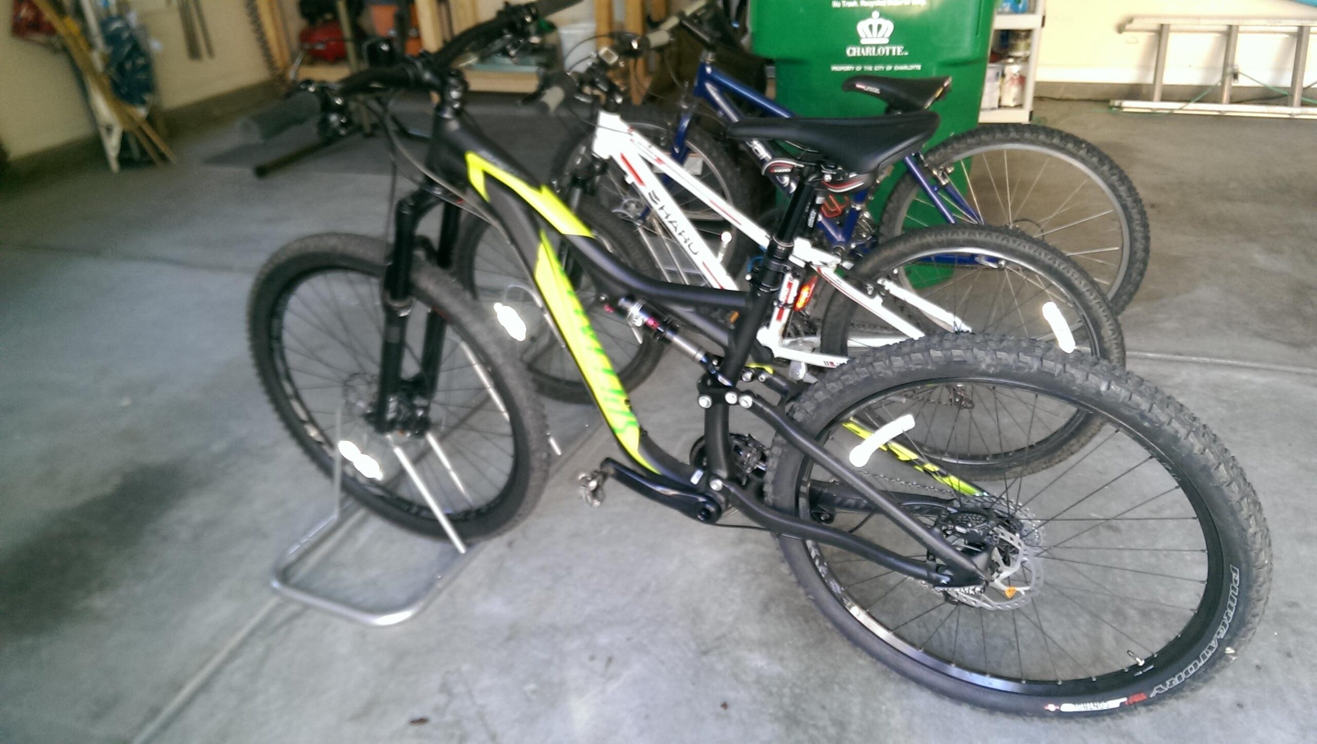 Specialized Stumpjumper FSR Comp EVO: A close-up view of two bicycles in a garage setting. The foreground features a black and lime green mountain bike on a bike stand, while a white mountain bike is partially visible behind it. The background shows additional bicycles and storage items, with a concrete floor and garage environment.