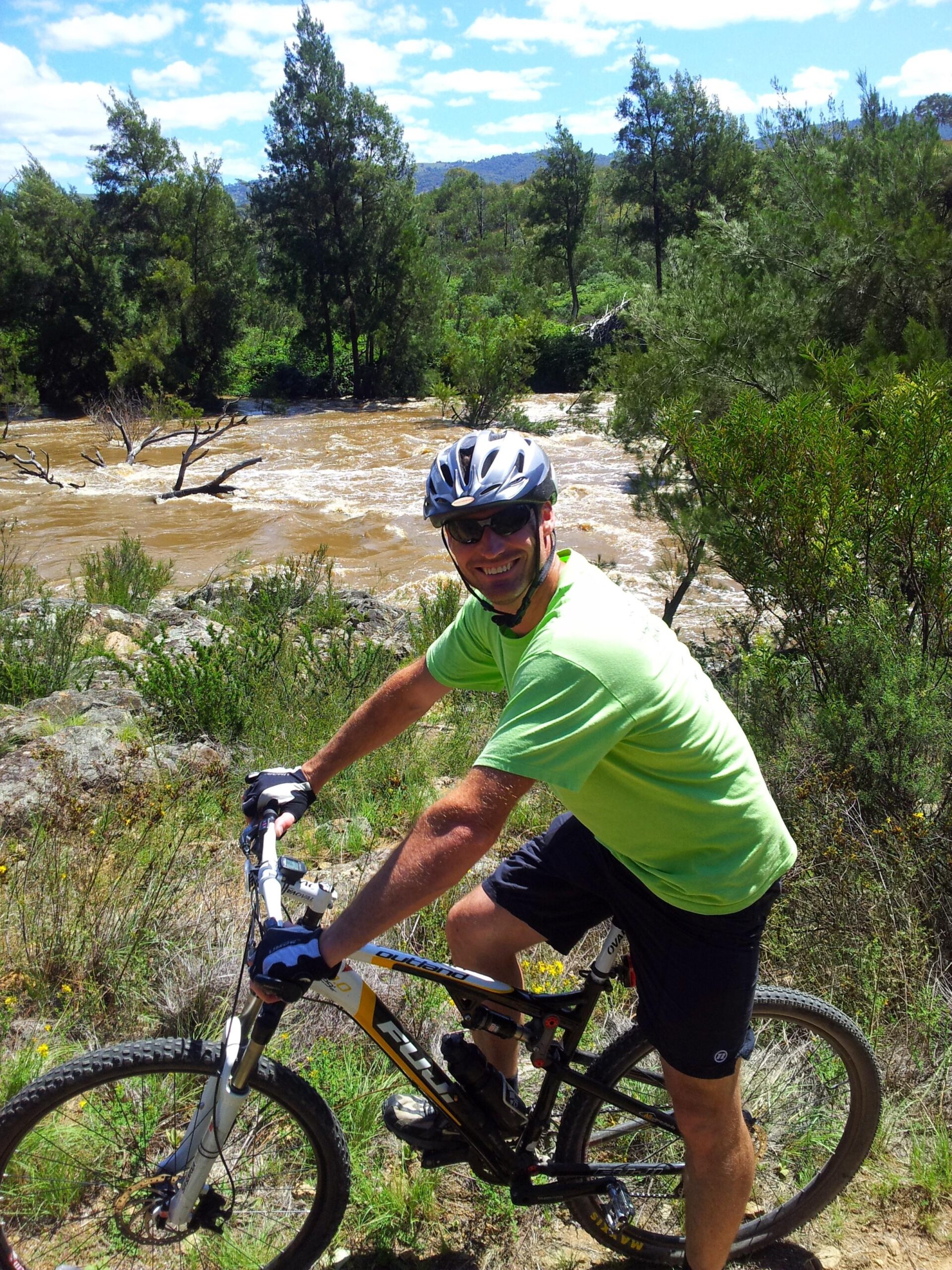 Fuji Outland 29 1.1 D: A man in a bright green shirt and cycling gear is smiling while riding a mountain bike beside a river with muddy water, surrounded by lush greenery and trees under a partly cloudy sky.