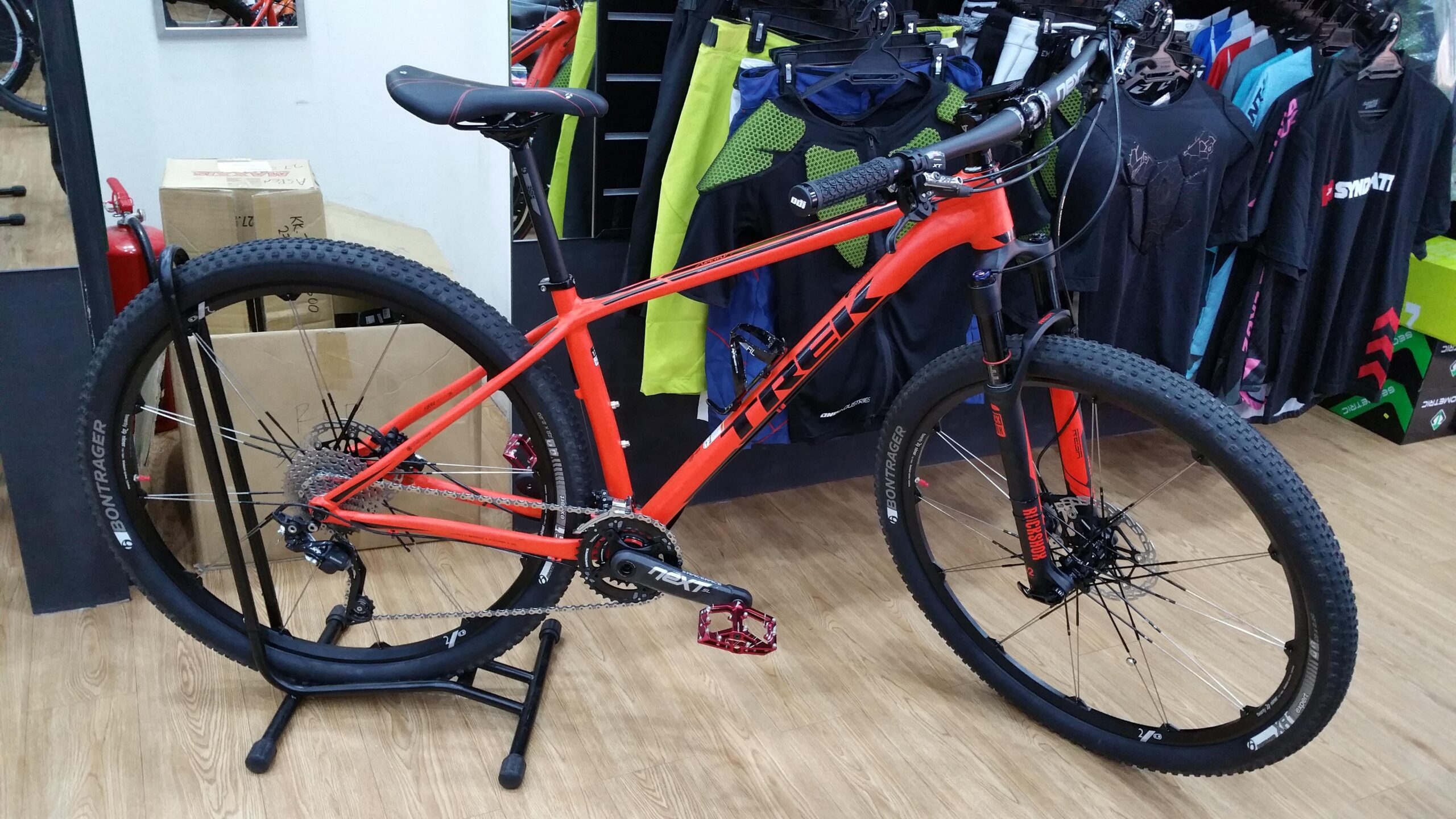 Trek Superfly 7: A bright orange Trek mountain bike displayed in a shop, featuring a sturdy frame, thick tires, and a detailed gear system. The bike is positioned on a stand with various cycling apparel hanging on the wall in the background.