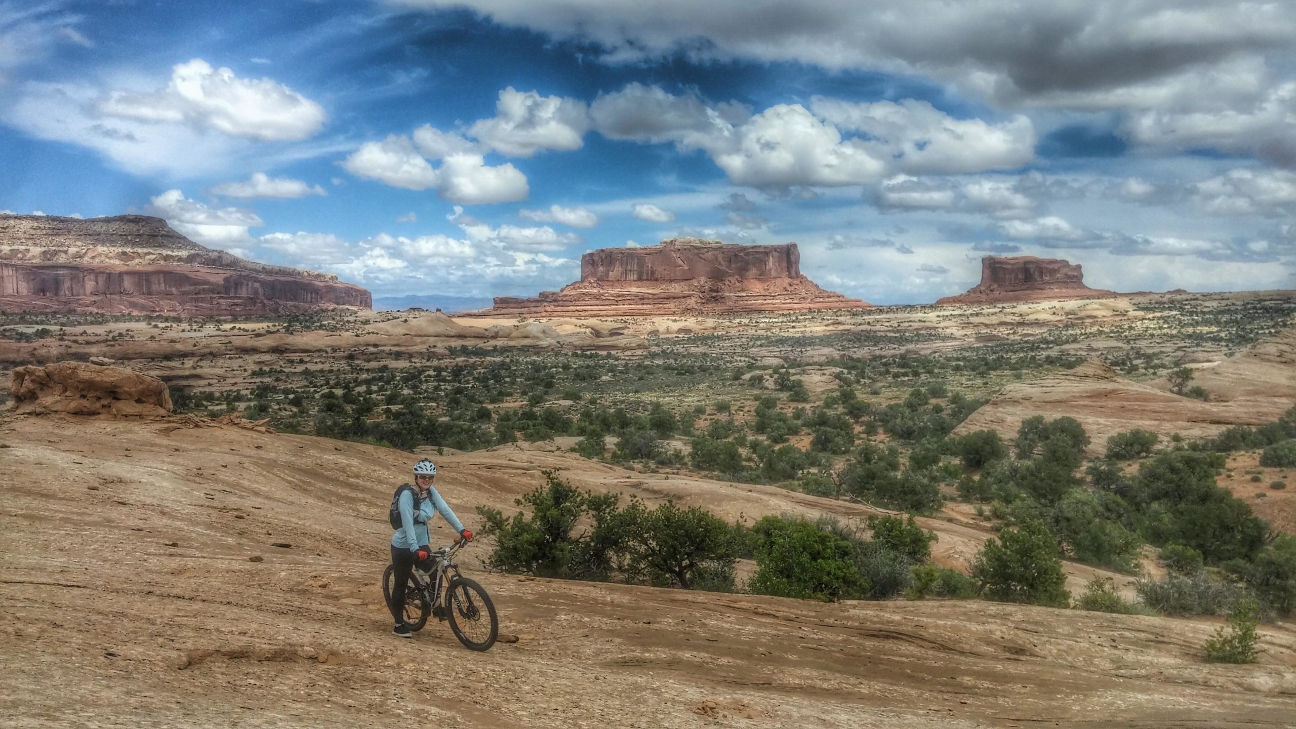 A cyclist on a mountain bike pauses on a rocky terrain, surrounded by stunning desert landscapes. In the background, dramatic mesas and plateaus rise against a cloudy blue sky, while clusters of greenery dot the rocky ground. Navajo Rocks mountain bike trail.