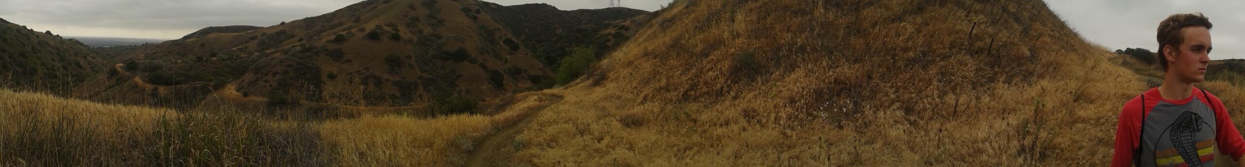 A panoramic view of a hiking trail winding through grassy hills under a cloudy sky, with a person standing to the right in casual clothing. The landscape features rolling hills with patches of greenery and dry grass. Turnbull Canyon mountain bike trail.