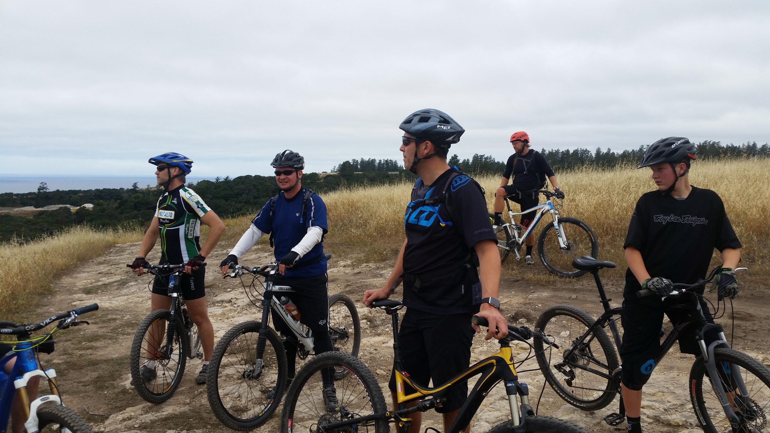 A group of six mountain bikers on a trail, standing next to their bikes. They are wearing helmets and cycling gear, surrounded by grass and trees, with a cloudy sky overhead and a view of distant hills in the background. Wilder Ranch State Park mountain bike trail.
