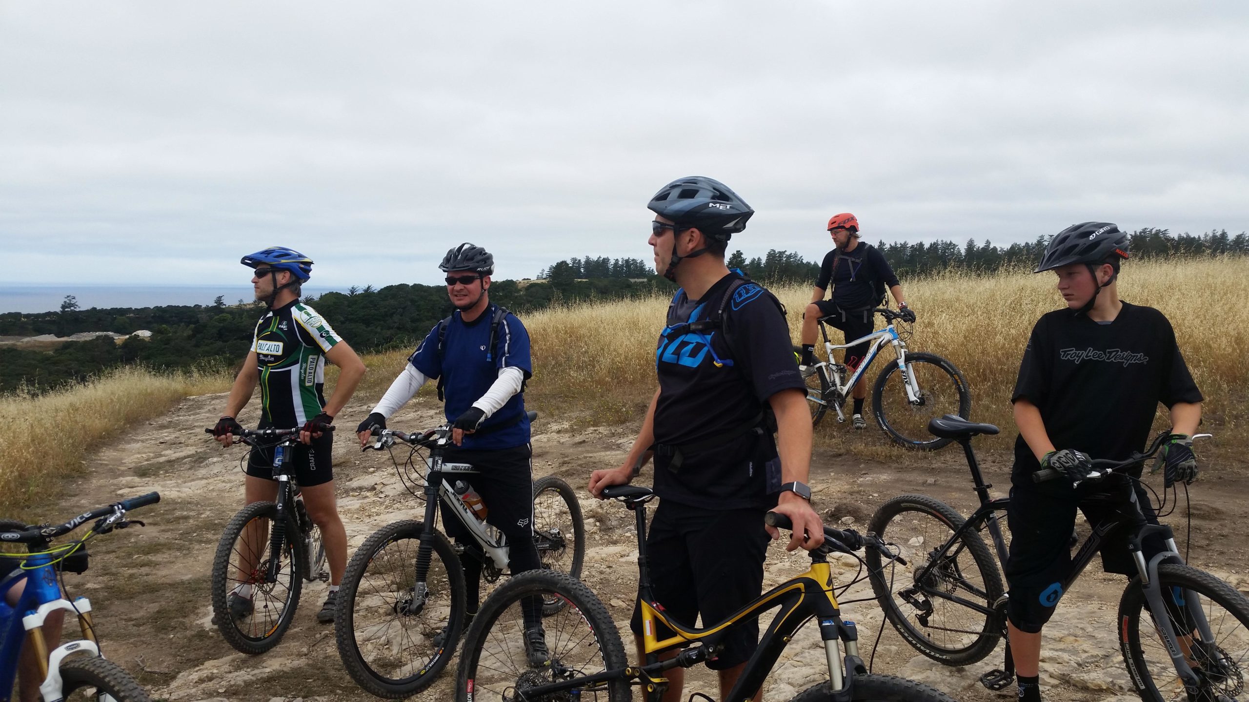 A group of six mountain bikers in helmets and cycling gear are gathered on a dirt trail surrounded by tall grass and trees. They are standing next to their bikes, engaged in conversation, with a cloudy sky and a view of the landscape in the background. Wilder Ranch State Park mountain bike trail.