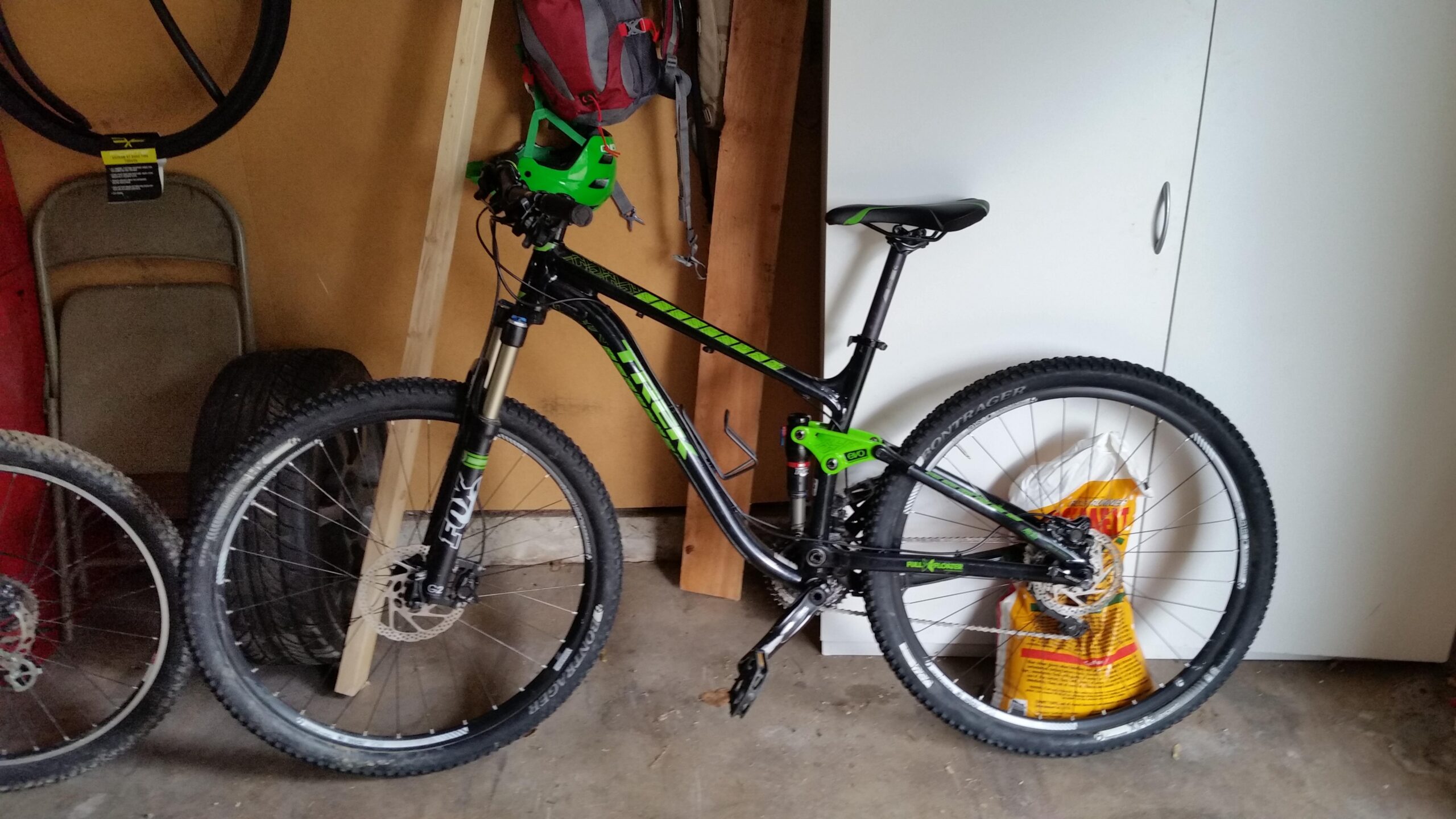 Trek Fuel EX 7 29er: A black and green mountain bike is leaning against a wall in a garage. The bike has large tires, a prominent suspension fork, and a sleek design. In the background, there is a stack of tires and some tools, along with a red backpack hanging on the wall. A bag of soil or mulch is resting against the bike.