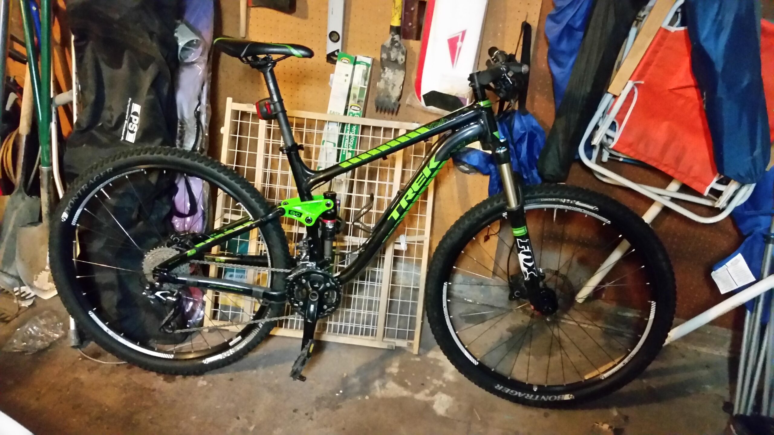 Trek Fuel EX 7 29er: A black and green mountain bike leaning against a wall in a garage, surrounded by various tools and equipment. The bike features wide tires, front suspension, and a visible brand name.